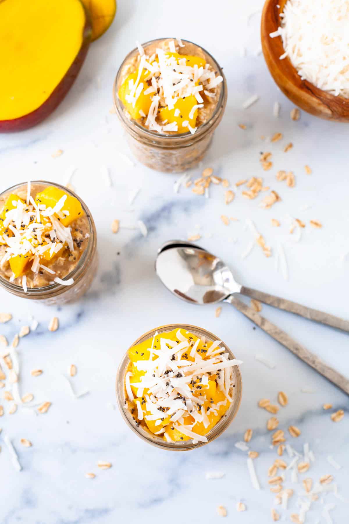 overhead. 3 mason jars with overnight oats. mango topping. mango slices. coconut shreds. raw oats. two spoons. 
