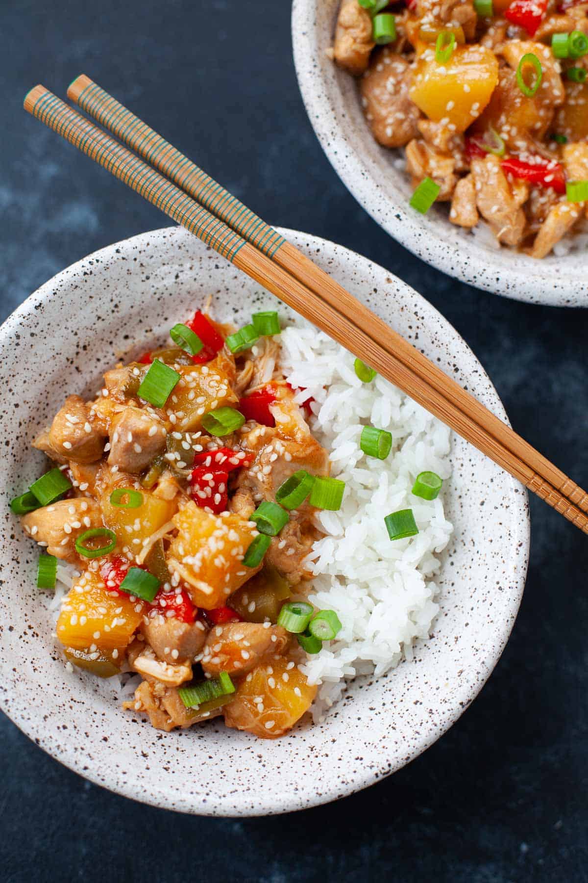 sweet and sour chicken over a bed of white rice with green onion in a grey speckled bowl with wood chopsticks