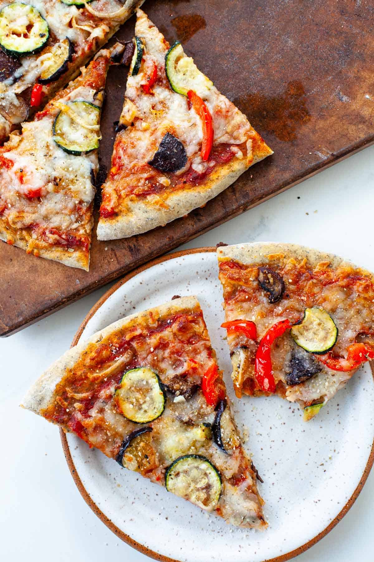 roasted vegetable pizza slices on white speckled plate and pizza stone with other pizza slices on it