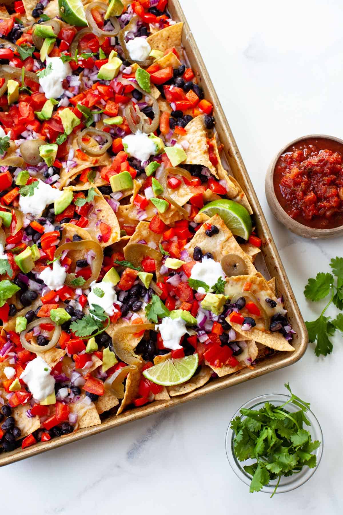 loaded vegetarian nachos on gold sheet pan with bowls of cilantro and salsa on the side