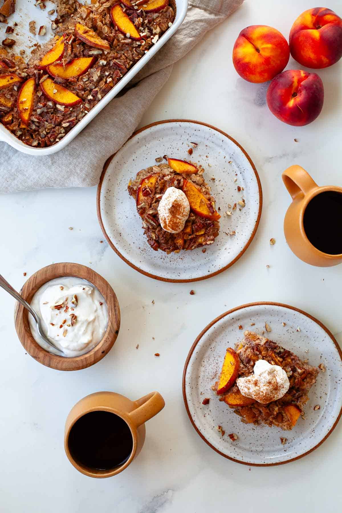 two white speckled plates with peach baked oatmeal beside two orange coffee mugs, a wooden bowl filled with yogurt, peaches, and a baking dish with peach baked oatmeal