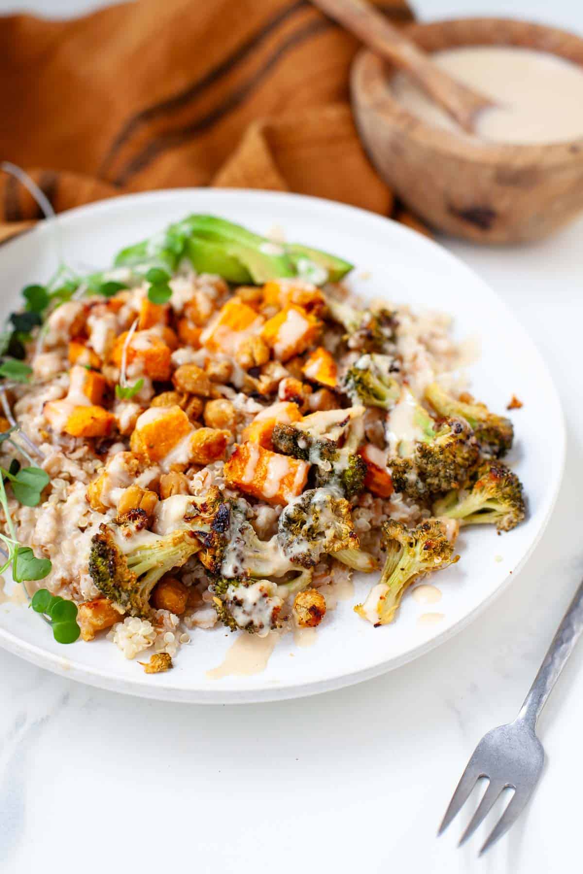 Ancient grains bowl with quinoa, farro, sweet potato, chickpeas, broccoli and avocado served in white bowl and drizzled with tahini sauce.