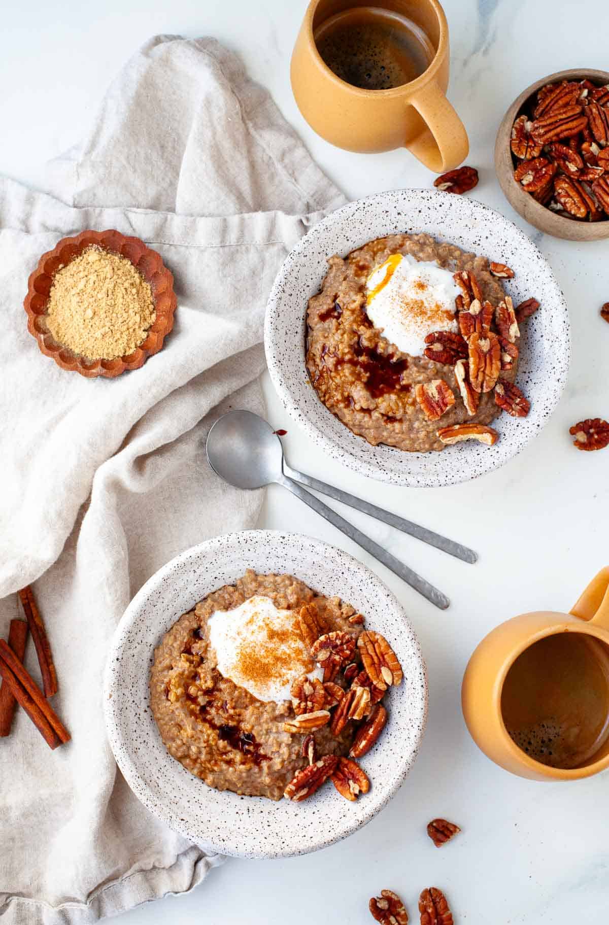 Two white speckled bowls filled with Gingerbread Oatmeal topped with yogurt, pecans and cinnamon, beside two orange coffee mugs, a small bowl of pecans, and a kitchen dish towel