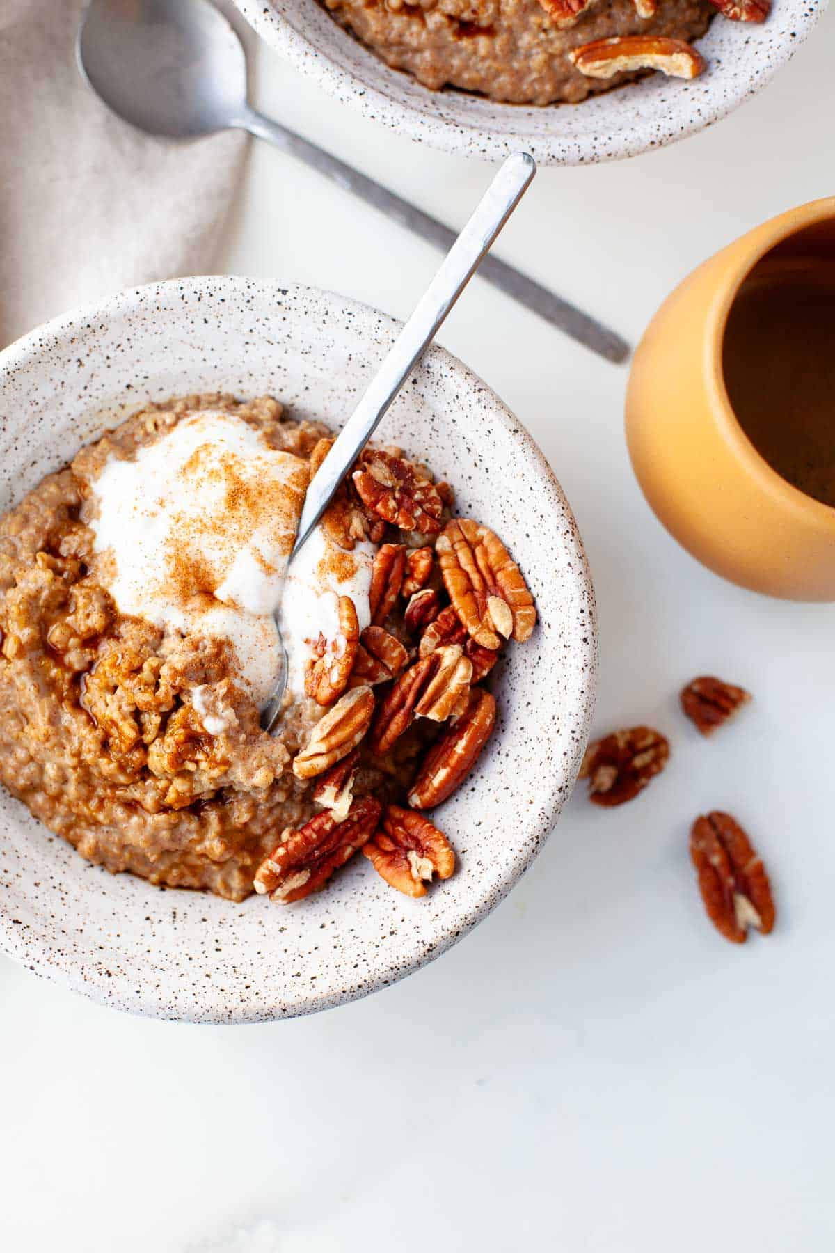 Gingerbread Oatmeal in a white speckled bowl garnished with pecans, yogurt, and cinnamon, with a silver spoon and tan coffee mug