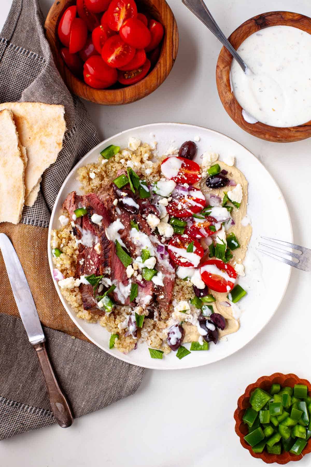 steak grain bowl surrounded by cherry tomatoes, herb yogurt dressing, chopped bell peppers, and pita bread.